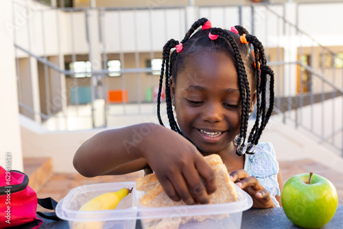African american schoolgirl at table and having healthy lunch with apple outside elementary school