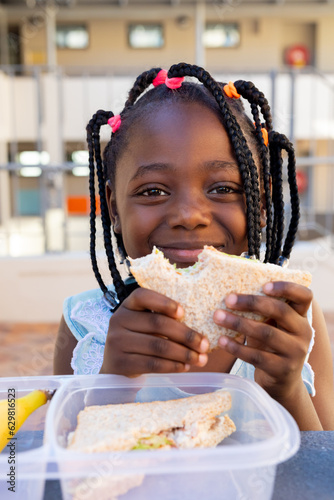 African american schoolgirl at table and having healthy lunch with sandwich at elementary school