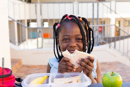 African american schoolgirl at table and having healthy lunch with sandwich at elementary school