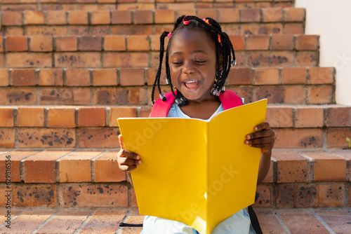 Happy african american schoolgirl sitting on stairs and reading book at elementary school