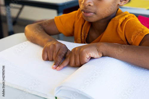 Focused biracial schoolboy reading braille with hands in classroom