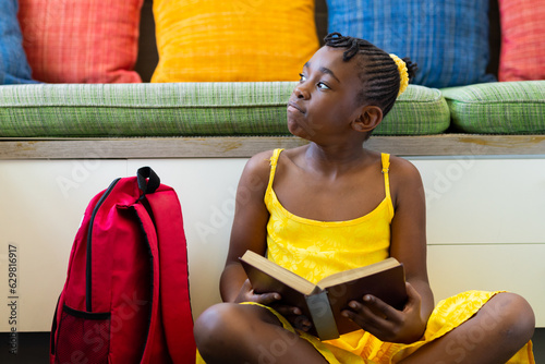 Thoughtful african american schoolgirl reading book over couch with colourful pillows