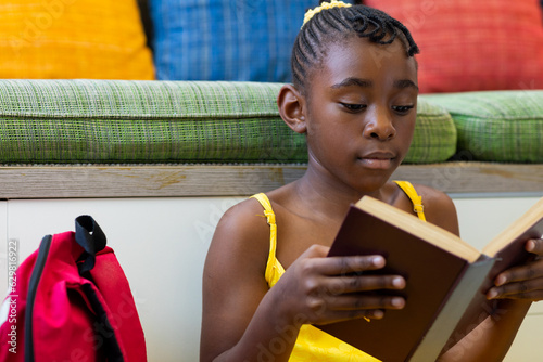 Focused african american schoolgirl reading book over couch with colourful pillows