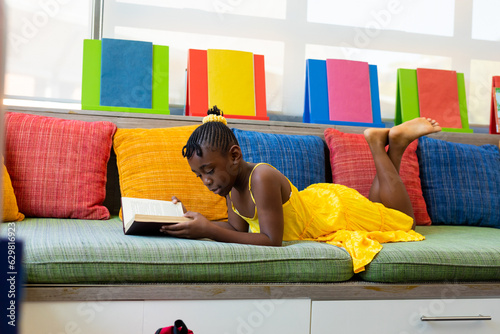 Focused african american schoolgirl reading book over couch with colourful pillows