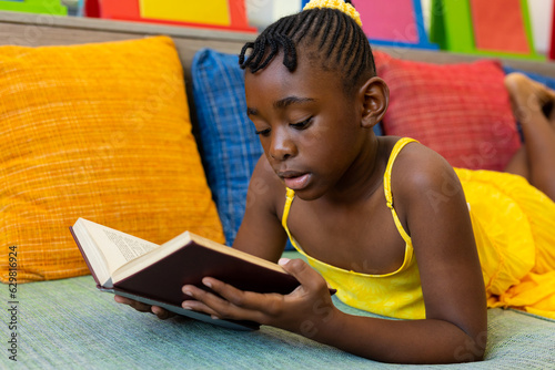 Focused african american schoolgirl reading book on couch with colourful pillows