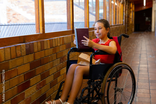 Happy caucasian schoolgirl sitting in wheelchair and reading book at school corridor