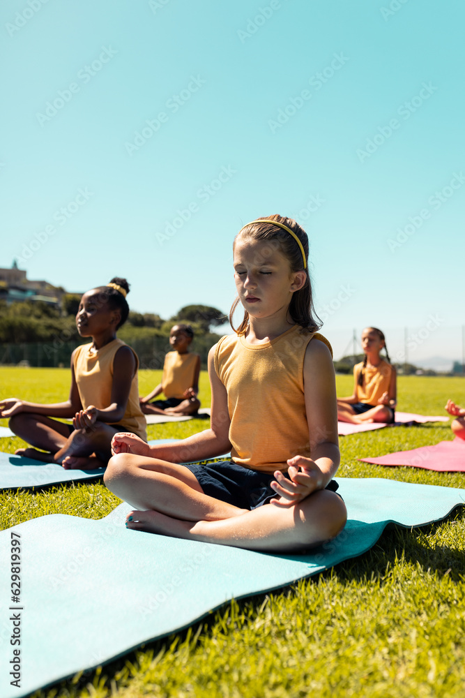 Fototapeta premium Diverse schoolgirls practicing yoga and meditating in sports field at elementary school, copy space