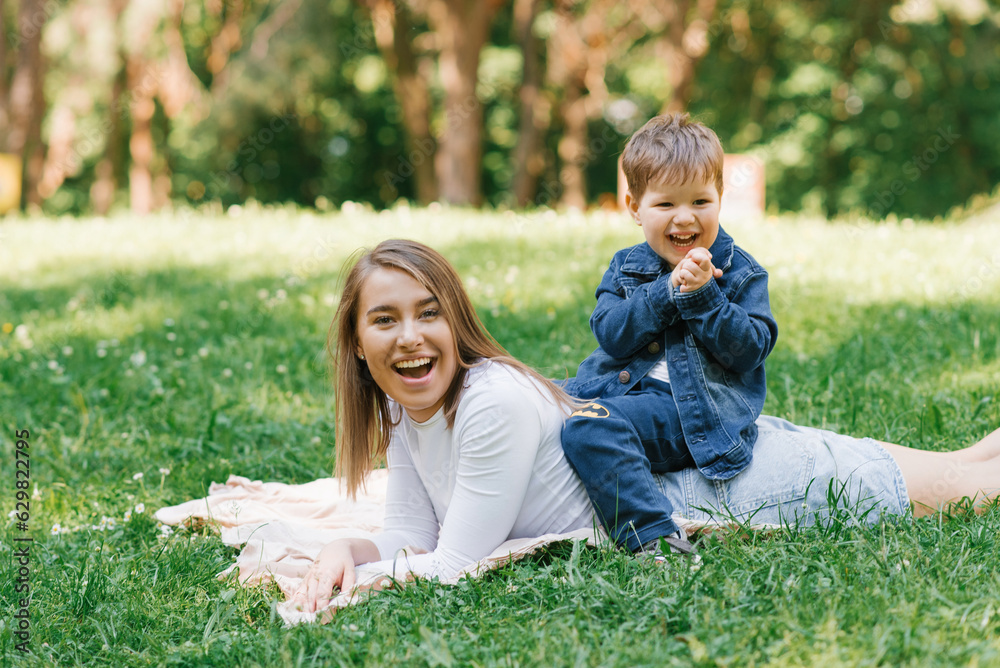 Young Caucasian mother and her young son lie on a blanket on the grass in the park in summer and relax outdoors