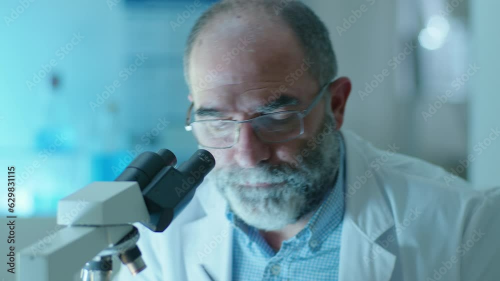 Senior male scientist with gray hair and beard wearing white coat and eyeglasses looking into microscope and writing down notes during research in laboratory Senior male scientist with gray hair and beard wearing white coat and eyeglasses looking into microscope and writing down notes during research in laboratory