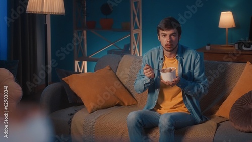 A man sits on a sofa in the living room and eats ice cream with a spoon from a bucket. A man with a full spoonful of ice cream looks straight ahead in surprise.