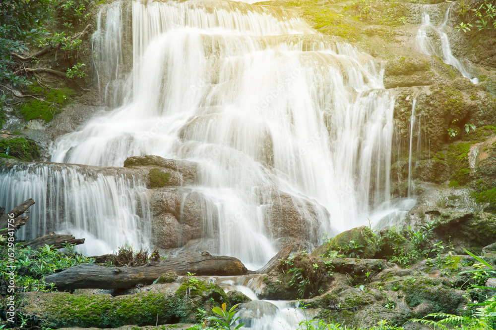 Fototapeta premium Natural waterfall view flowing through the rocky mountain with tropical forest and moss in sun light effect and dark green tone