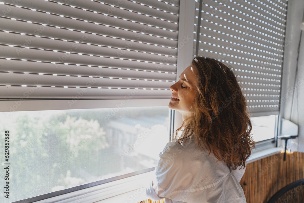 Young woman opening roller blind shutters on the balcony on a summer day
