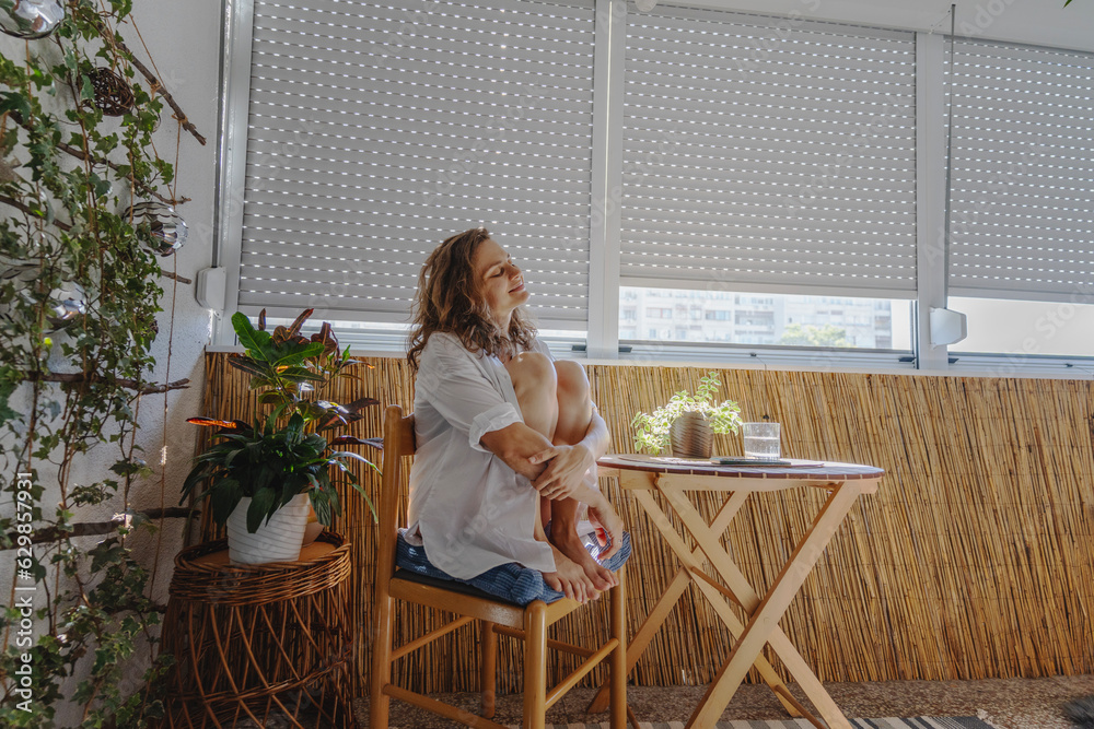 Young caucasian woman in a white shirt sitting on a balcony with roller shutters. Sun protection and rising temperature concept