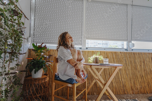 Obraz na płótnie Young caucasian woman in a white shirt sitting on a balcony with roller shutters. Sun protection and rising temperature concept w salonie