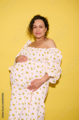 Pregnant woman in late pregnancy gently strokes her big belly, looking confidently at camera, isolated yellow background photo