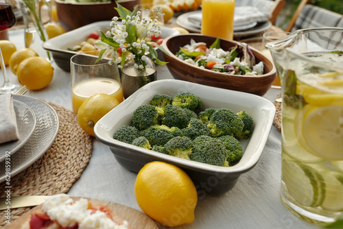 Container with steamed or fresh broccoli surrounded by lemons and other food standing on served table among variety of homemade stuff