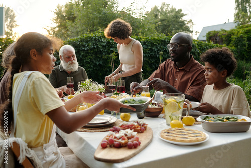 Modern family of siblings, their intercultural parents and grandparents sitting by dinner table served with homemade food and drinks