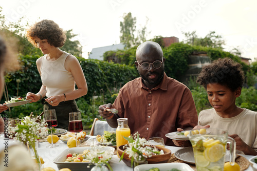 Young African American man and his son putting homemade food on plates while sitting by served table during outdoor family dinner