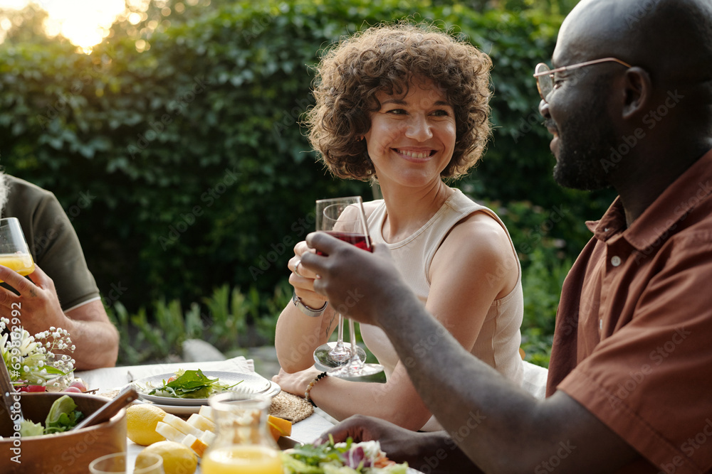 Fototapeta premium Happy young brunette woman toasting with her husband with glasses of wine while both sitting by dinner table and enjoying outdoor gathering