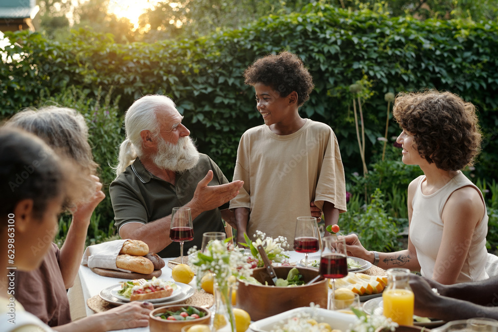 Cute happy African American boy looking at his grandfather during talk while standing in front of served table with his family having dinner Cute happy African American boy looking at his grandfather during talk while standing in front of served table with his family having dinner
