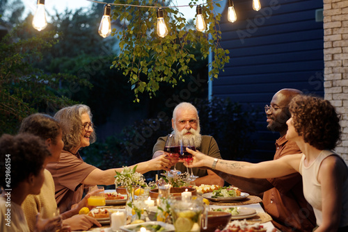 Side view of large intercultural family of six clinking with glasses of red wine over table served with homemade food during outdoor dinner