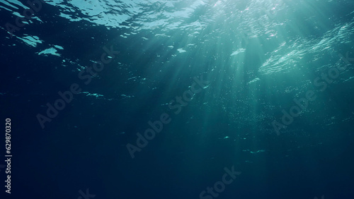 View of sun light from blue abyss, slow motion. Light filters down through blue water. Underwater sun rays in depth ocean. Underwater sun light shine under deep water with ripples on waves surface
