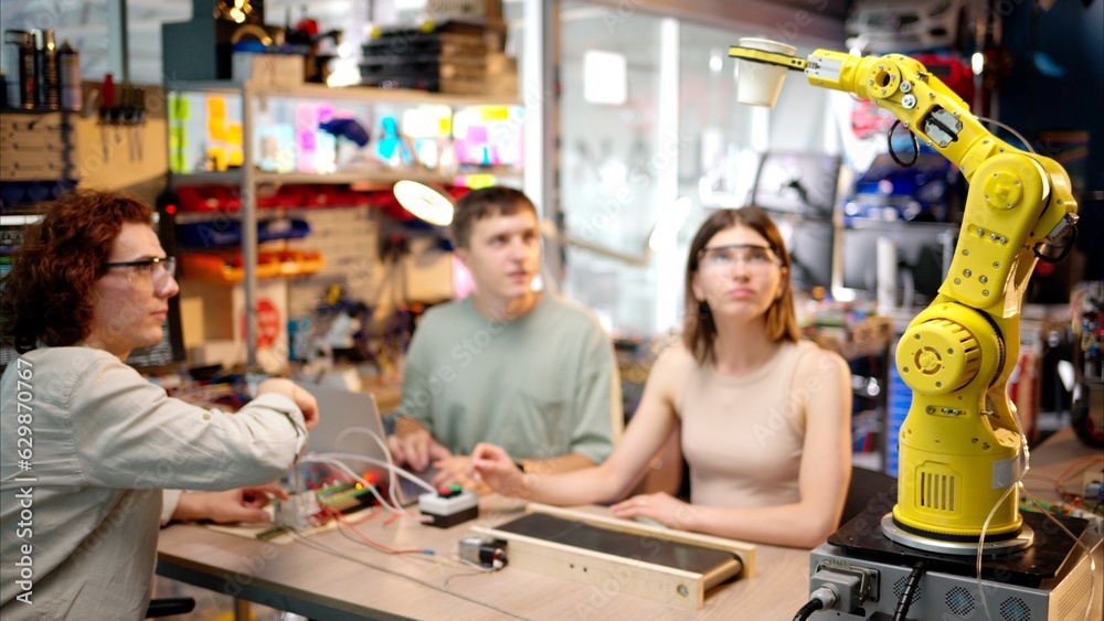 Three young happy engineers fixing an yellow robotic arm in the workshop, computer programming Three young happy engineers fixing an yellow robotic arm in the workshop, computer programming