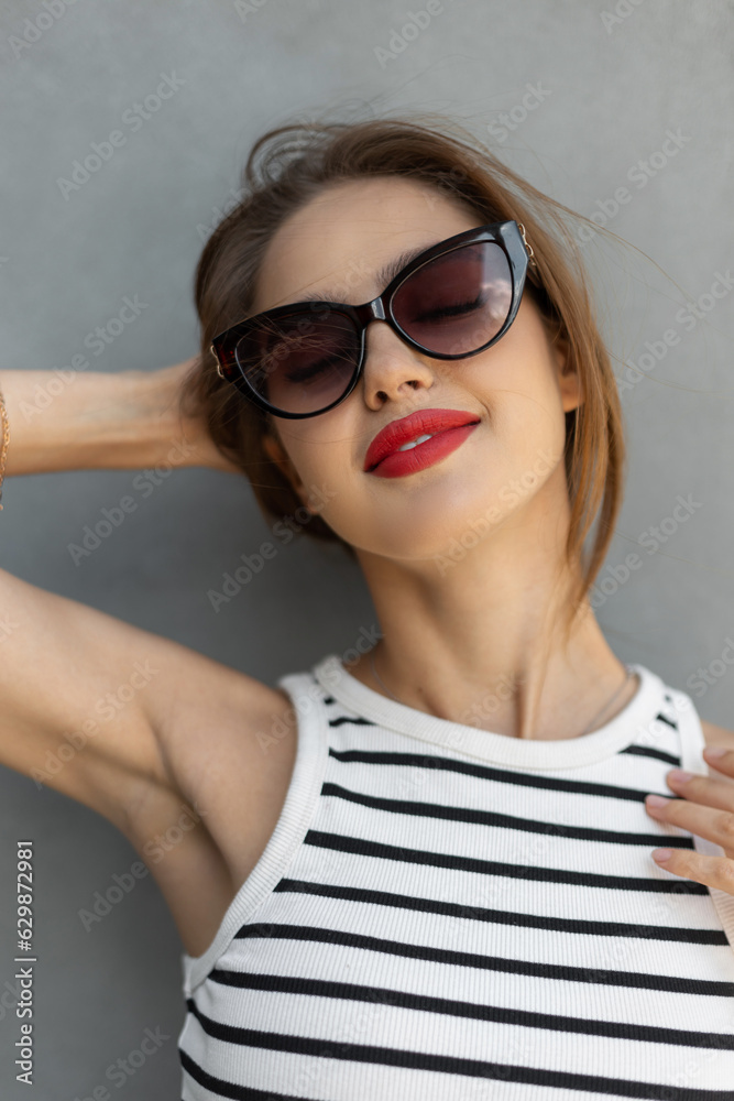 Happy beautiful young fresh woman with a smile with vintage fashion sunglasses stands near a gray wall on the street Happy beautiful young fresh woman with a smile with vintage fashion sunglasses stands near a gray wall on the street