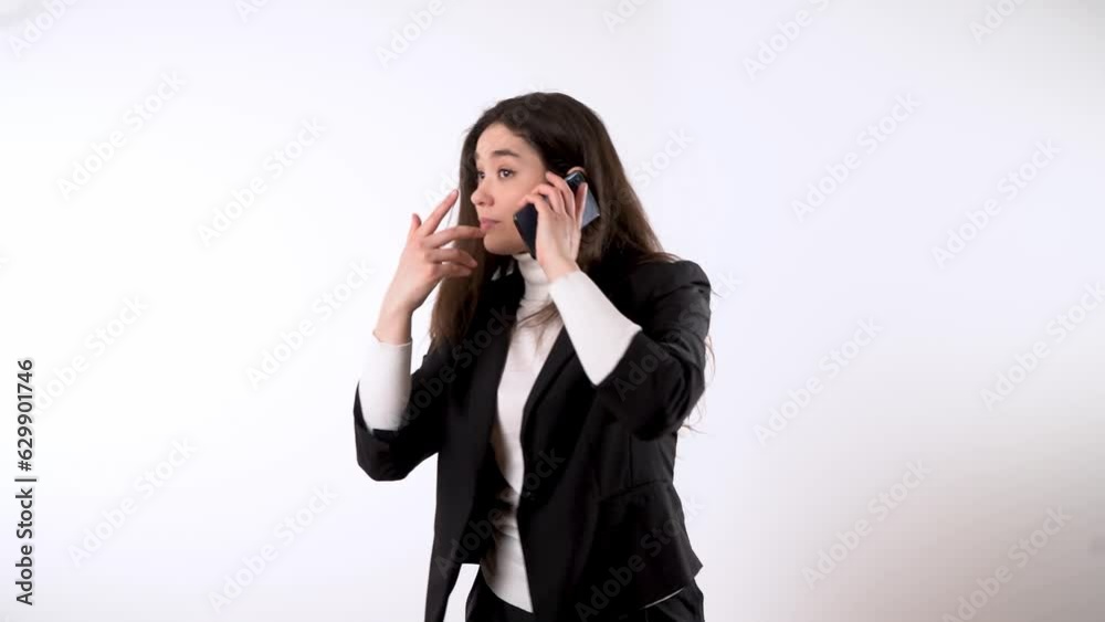 Nervous young business lady in formal suit with dark hair against white background talking on her smartphone while angrily gesturing someone to keep silent Nervous young business lady in formal suit with dark hair against white background talking on her smartphone while angrily gesturing someone to keep silent