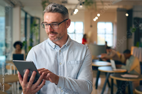 Mature Businessman Standing In Busy Office Using Digital Tablet