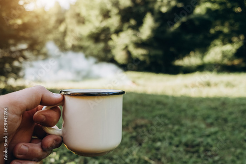 Close up of unknown young caucasian man holding hot coffee near beautiful bonfire in summer morning forest. Hiking guy enjoying warm nature. Wanderlust adventures in outdoor living lifestyle, hiking