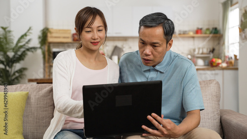 asian smiling daughter and confused elderly father sitting together with a laptop computer on living room sofa at home. she teaches him how to surf the internet with gestures © PR Image Factory