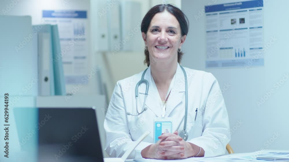 Mature female general practitioner in white coat with stethoscope over neck sitting at workplace in clinic, looking at camera with a happy smile. Medium shot, video portrait