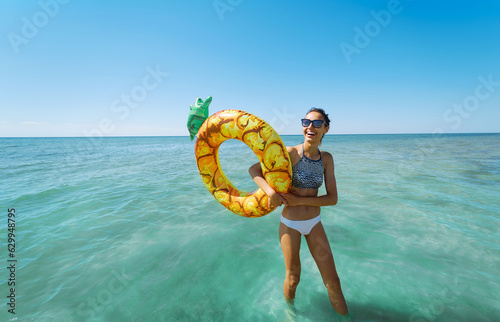 Happy vacation woman in swimwear standing in the sea with inflatable pineapple float ring. Carefree beach holidays and feel the warmth of the sun