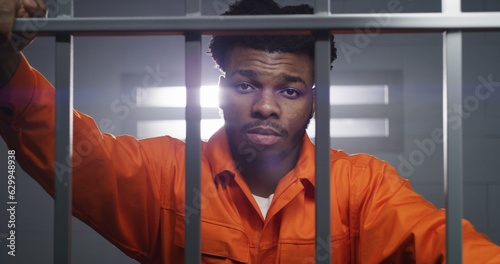 African American man in orange uniform looks at camera and kneads neck in prison cell. Prisoner serves imprisonment term in jail. Criminal in correctional facility or detention center. Portrait view.