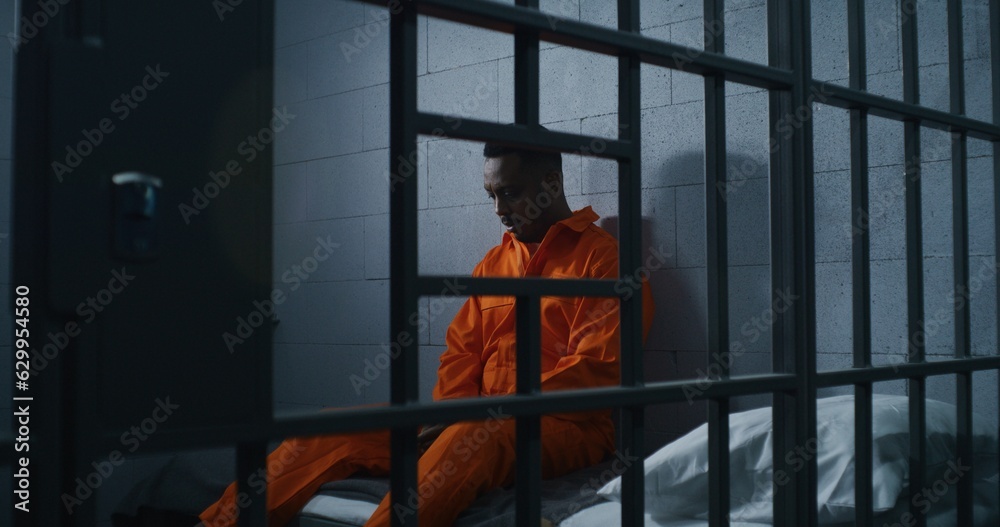 Guilty African American man in orange uniform sits on prison bed and thinks about freedom. Gloomy criminal in correctional facility or detention center. Prisoner serves imprisonment term in jail cell. Guilty African American man in orange uniform sits on prison bed and thinks about freedom. Gloomy criminal in correctional facility or detention center. Prisoner serves imprisonment term in jail cell.