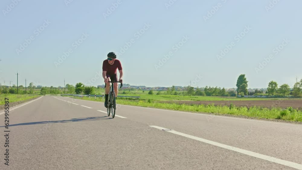 Young professional cyclist in sportswear riding down beautiful countryside road between fields. Athletic adult man promoting active lifestyle while practising strength workout on sunny summer day. Young professional cyclist in sportswear riding down beautiful countryside road between fields. Athletic adult man promoting active lifestyle while practising strength workout on sunny summer day.