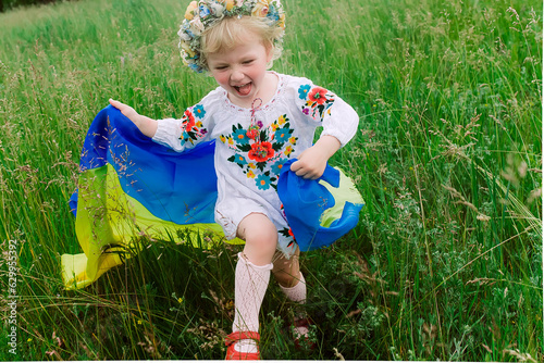 flag of Ukraine is on the shoulders of a little Ukrainian girl. Happy Ukrainian child in free Ukraine without war runs along a rural road in a field of farm soybean. International Mother Language Day.