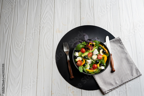 Greek salad in a deep black bowl, knife and fork on a black round stand on a light wooden background.