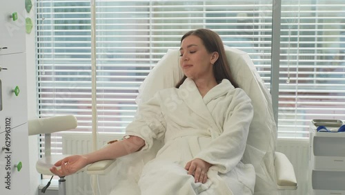 Tracking shot portrait of pretty young woman patient in white bathrobe with IV catheter in vein receiving vitamin treatment in beauty salon lying on comfy medical armchair. Shooting in slow motion.
