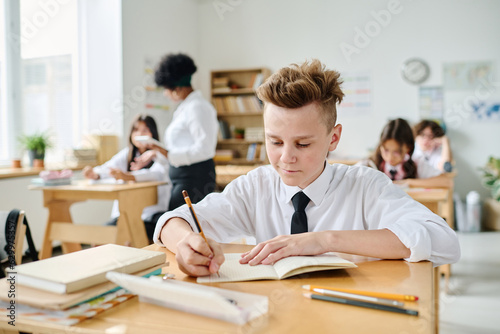 Schoolboy making notes in notebook while sitting at desk during lesson