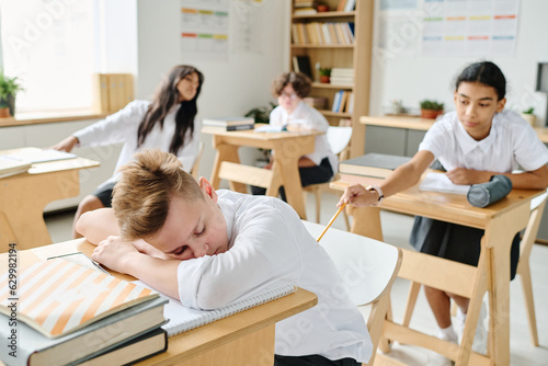 Schoolboy sleeping at desk at lesson while his classmate waking him up