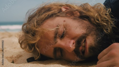 Male person lying on sand beach suffering from hangover. Homeless man in depression listening to wave. Peaceful noise of ocean. Sun stroke. Tired smile of solo traveler. Stray tramp sleeping outdoors. photo