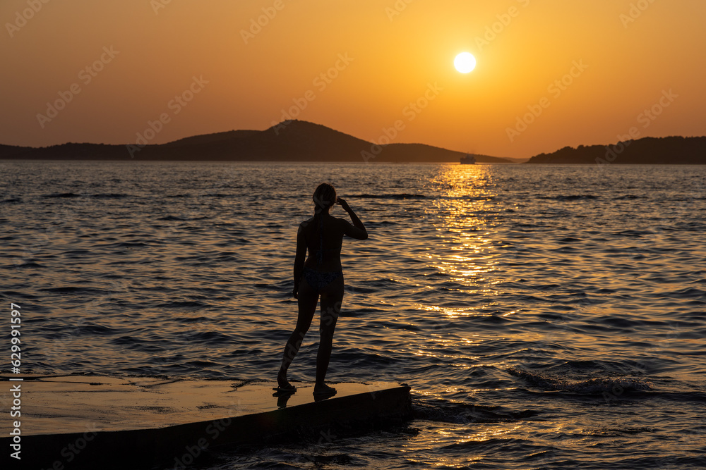 A young woman is standing on a pier, looking at the sunset on the Adriatic Sea in Croatia.