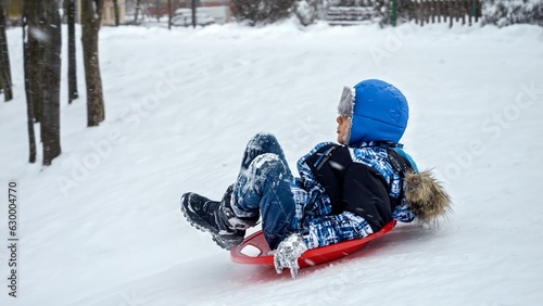 Cheerful boy riding down the snowy hill on plastic sleds during snowfall. Concept of winter holiday, children having fun and playing outdoors in the snow, Christmas vacation.