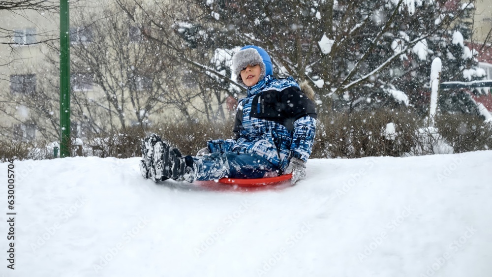 Cheerful smiling boy riding down the snowy hill on red plastic sleds during snowfall. Cheerful smiling boy riding down the snowy hill on red plastic sleds during snowfall.