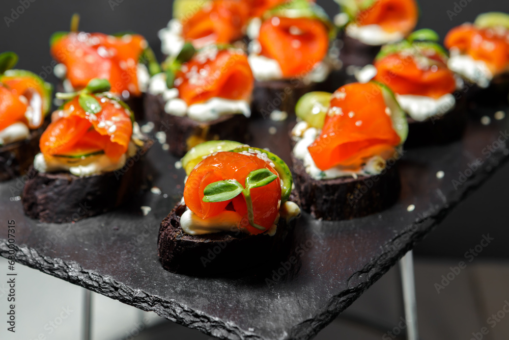 Canapes of black bread, red fish and cucumbers, seasoned with sauce and sprinkled with sesame seeds, lie on a tray. A fragment of the serving. Canapes of black bread, red fish and cucumbers, seasoned with sauce and sprinkled with sesame seeds, lie on a tray. A fragment of the serving.
