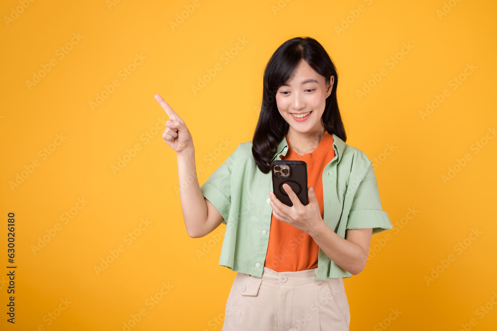mobile apps with enchanting young Asian woman 30s, wearing orange shirt, happily pointing finger gesture to free space while holding smartphone isolated on yellow background. app smartphone concept. mobile apps with enchanting young Asian woman 30s, wearing orange shirt, happily pointing finger gesture to free space while holding smartphone isolated on yellow background. app smartphone concept.