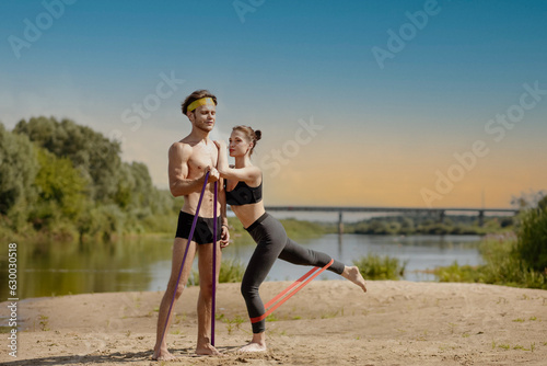 Fototapeta Naklejka Na Ścianę i Meble -  Sports couple exercising with resistance band outdoors. Handsome man and sexy woman exercising on the coast. Active summer vacation exercises for a strong muscular body.