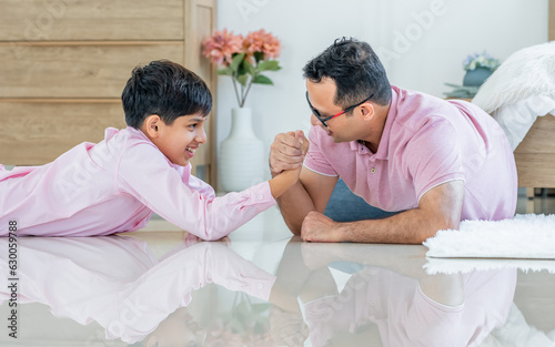 Indian happy father and son playing together, laying down on floor, doing arm wrestle with amusement and fun while staying in living room at cozy home in weekend. Education, Family, Activity Concept.